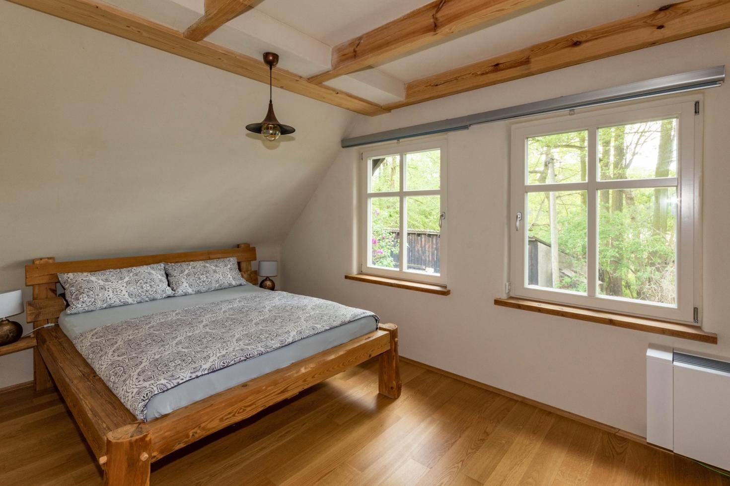 Schlafzimmer eines Naturapartments mit großem Holzbett, sichtbaren Deckenbalken und Fenstern mit Blick auf die umgebende Natur.